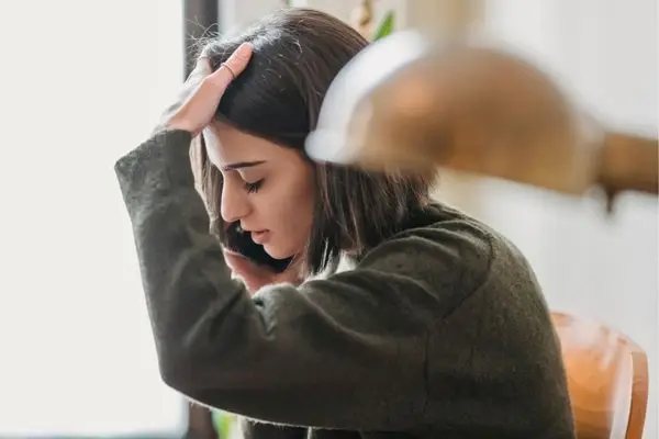 A young woman sits indoors, holding a phone to her ear with one hand on her head, looking stressed during a somatic therapy session